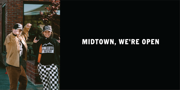 Two people in casual clothing with a black screen displaying 'MIDTOWN, WE'RE OPEN'.