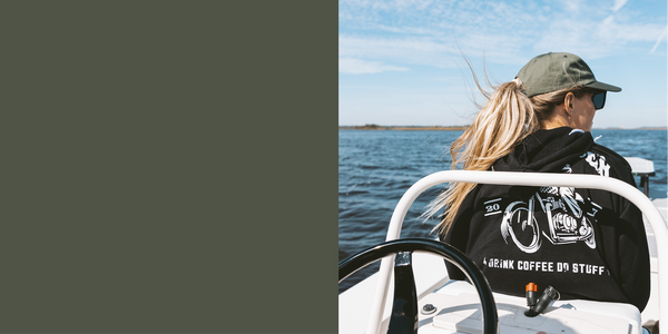 The photo shows the back of a blonde woman's head, hair in a ponytail blowing in the wind, sitting on a boat. The blue sky and blue water are visible in the background behind her, and she's wearing a black Drink Coffee Do Stuff sweatshirt with an army green dad hat.